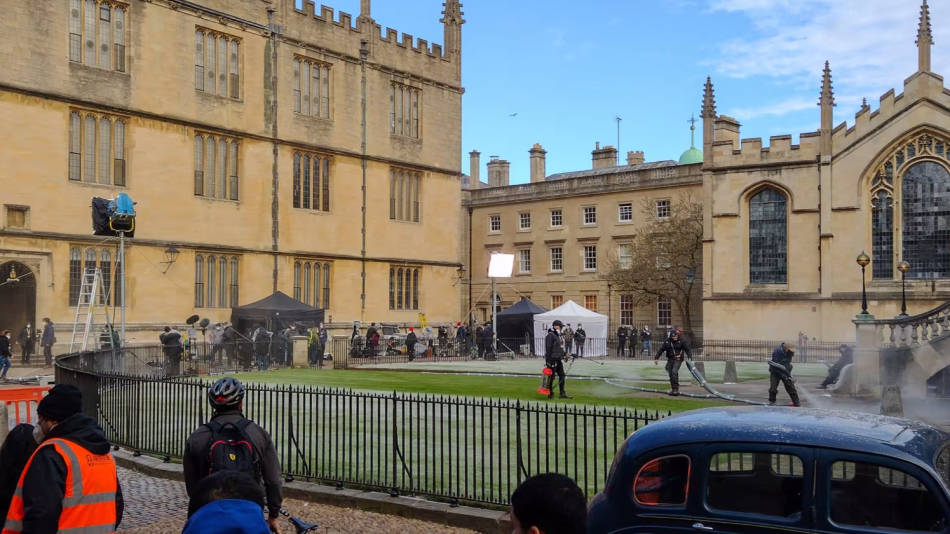 A wide view of a historic Oxford college courtyard on a bright day. Large Gothic stone buildings with tall windows and decorative spires surround a grassy quadrangle. The area appears to be hosting a film or television production, with lighting rigs, tents, crew members, and equipment set up around the lawn. Several workers are spraying water or creating mist near a stone staircase on the right. In the foreground, people stand behind a black iron fence, and a dark vintage-style car is parked beside the cour