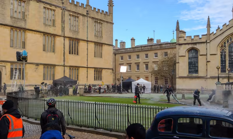 A wide view of a historic Oxford college courtyard on a bright day. Large Gothic stone buildings with tall windows and decorative spires surround a grassy quadrangle. The area appears to be hosting a film or television production, with lighting rigs, tents, crew members, and equipment set up around the lawn. Several workers are spraying water or creating mist near a stone staircase on the right. In the foreground, people stand behind a black iron fence, and a dark vintage-style car is parked beside the cour